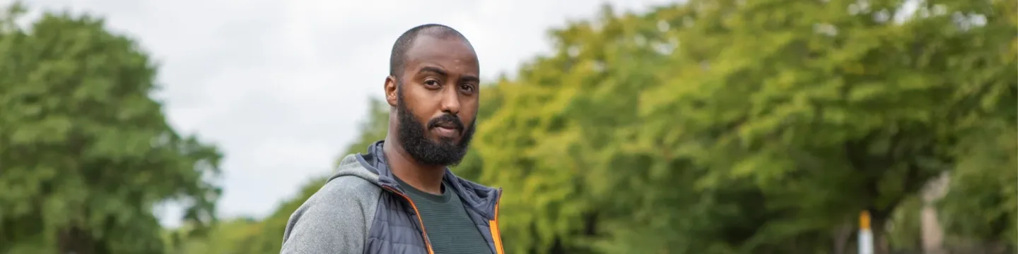 A professional who works with homeless Londoners, with a short beard and shaved head, wearing a green shirt and a gray jacket with an orange zipper, stands outdoors with trees and a cloudy sky in the background.