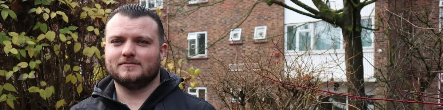 A man wearing a black jacket with slicked-back hair stands with trees and houses in the background.