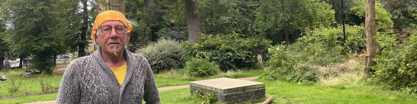 An elderly man wearing a yellow bandana stands in a graveyard, surrounded by gravestones and trees in the background.
