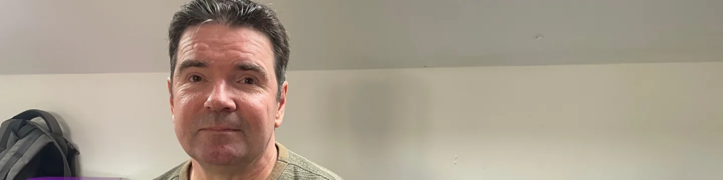 A man is sitting on a bed indoors, wearing a patterned sweater.