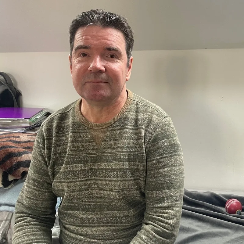A man is sitting on a bed indoors, wearing a patterned sweater.