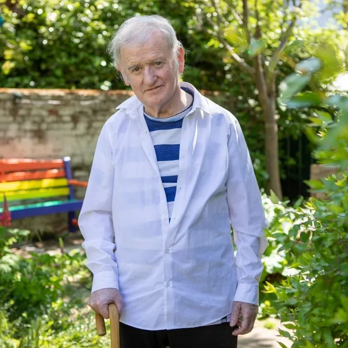 A elderly white man wearing a white shirt and with a small smile stands in a summery leafy garden, with his walking stick