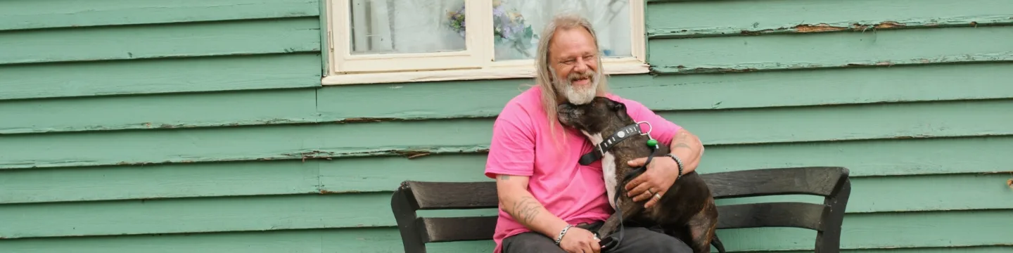 A man in a pink shirt relaxes on a bench, enjoying the company of his dog on his lap.
